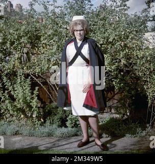 1960s, storica, in piedi fuori per la sua foto, una giovane infermiera femminile nella sua uniforme di abito a righe e grembiule bianco e sopra di esso, con un mantello nero, con chiusura a croce e fodera color marrone, Inghilterra, Regno Unito. Il mantello è stato progettato per essere aperto sul davanti. Sulla sua testa è una tradizionale cuffia da infermiera e sta indossando gli occhiali che erano alla moda negli anni '50s e '60s, noti come occhiali Cat Eye. Foto Stock