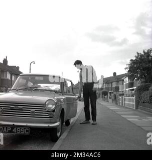 1960s, storico, un uomo in piedi fuori sul marciapiede in una strada suburbana che apre la porta della sua auto, una Austin A40 farina, una piccola economia auto dell'epoca, che era in produzione tra il 1958 e il 1967. Il suo nome deriva dal designer dell'auto, lo studio 'Pinin farina' di Torino. Foto Stock