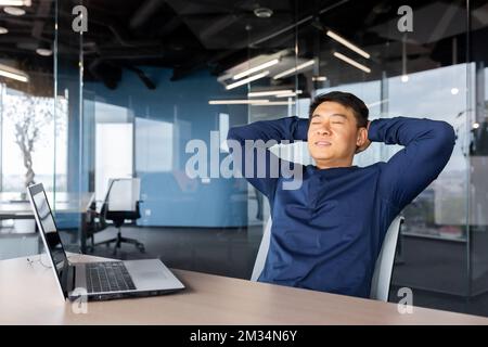 Un uomo d'affari di successo ha finito bene il lavoro, l'uomo asiatico ha messo le mani dietro la testa e riposarsi sulla sedia con gli occhi chiusi, il dipendente lavora in ufficio utilizzando un computer portatile al lavoro. Foto Stock