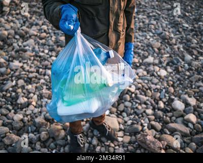 Processo di pulizia della spiaggia di pietra dai rifiuti di plastica. L'uomo tiene bottiglie di plastica nel sacchetto dei rifiuti. Concetto di inquinamento ambientale. Foto Stock