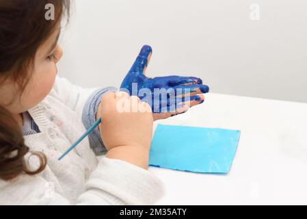 Bambino dipinge la sua mano con vernice blu e pennello diventando disordinato e divertirsi. Foto Stock