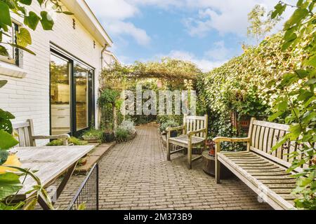 un'area esterna con panchine e piante sul lato della strada di fronte ad una casa bianca, circondata da un lussureggiante verde fogliame Foto Stock