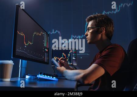 Vista laterale di un analista professionista giovane al tavolo di lavoro, analisi del grafico finanziario, quotazioni azionarie, prezzi delle azioni, trading online, verifica dei dati sul grafico della criptovaluta sullo schermo del computer, prendendo appunti Foto Stock