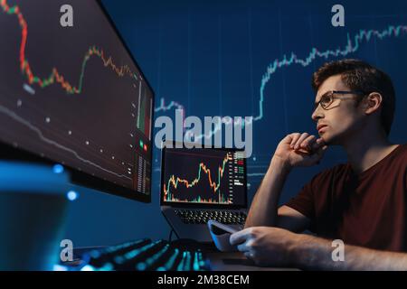 Serio commerciante criptato del tipo che guarda lo schermo con la tabella del candelabro, tenendo la penna ed il blocco note, facendo le note, analizzando i rischi di criptovaluta di commercio nelle condizioni della crisi finanziaria globale Foto Stock