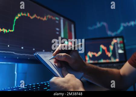 Nessuna immagine facciale di mani maschili scrivere numeri in blocco note dopo l'analisi del diagramma di mercato di valuta cripto al suo pc, prevedendo modelli di flusso di denaro, inventando una nuova strategia evitando rischi elevati Foto Stock