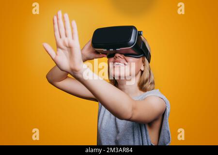 Donna attraente utilizzando occhiali di realtà virtuale, allungando il palmo, cercando di toccare qualcosa, isolato su sfondo giallo. Visore VR. Futuro Foto Stock