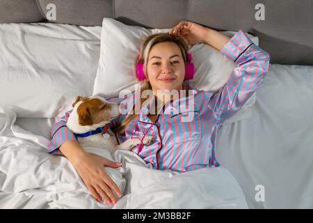 Donna spensierata in pigiama ascoltare musica sulle cuffie mentre si siede a letto con il suo amato animale domestico Foto Stock