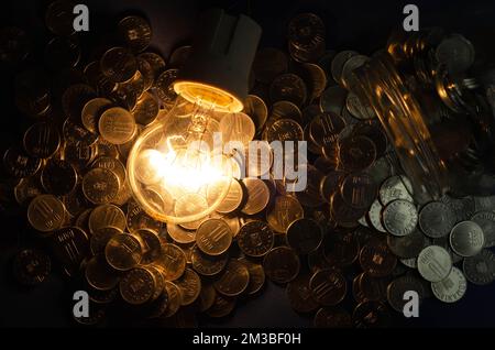 Concetto di consumo di elettricità, in questi tempi di crisi, rappresentato da una lampadina e monete in euro sparse, sfondo scuro Foto Stock