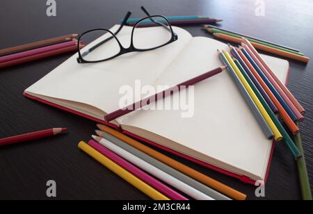 Occhiali neri con taccuino rosso bianco e matite colorate su tavolo di legno. Foto Stock