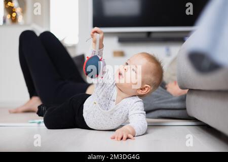 Momenti felici con la famiglia. Madre stesa comodamente sul tappeto dei bambini che guarda e sorveglia il suo bambino playinghis in salotto. Emozioni umane positive, sentimenti, gioia. Foto Stock