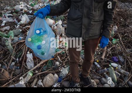 Processo di pulizia della spiaggia di pietra dai rifiuti di plastica. L'uomo tiene bottiglie di plastica nel sacchetto dei rifiuti. Concetto di inquinamento ambientale. Foto Stock
