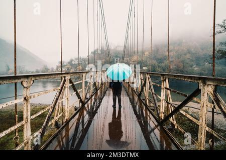 Ragazza con ombrello blu su ponte sospeso in ferro bagnato in caso di pioggia, vista posteriore. Foto Stock