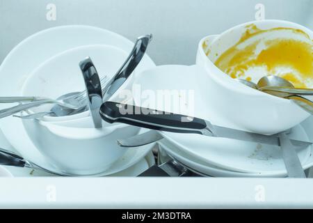 Primo piano di lavello bianco pieno di stoviglie bianche sporche, piatti, ciotole, coltelli, forchette e cucchiaio. Cucina, pulizie, pulizia, utensili, cucina per la casa Foto Stock