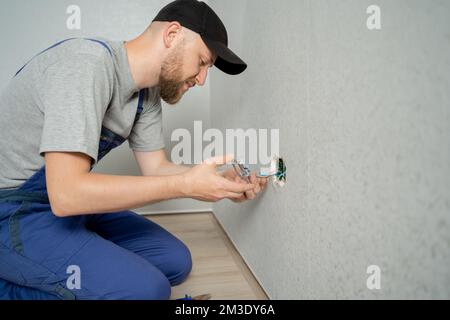 elettricista caucasico che tiene il cacciavite lavorando sulla spina elettrica su un impianto elettrico residenziale che installa la presa di alimentazione sulla parete grigia a casa Foto Stock