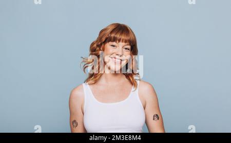 Ritratto di una giovane donna con capelli di zenzero sorridenti alla fotocamera. Donna con tatuaggi in piedi su uno sfondo blu in una canotta. Foto Stock