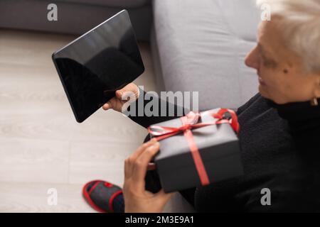 Sorridente donna caucasica di mezza età seduto sul divano in soggiorno con Internet wireless sul tablet, felice donna moderna senior rilassarsi sul divano a casa Foto Stock