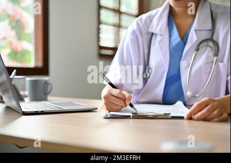 immagine ritagliata. Medico femminile asiatico intelligente millenario prendendo appunti o compilando la forma medica alla sua scrivania. Foto Stock