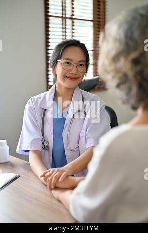 Una giovane e bella dottoressa asiatica tiene la mano di un paziente per confortare e rassicurare il paziente durante il controllo medico. Foto Stock