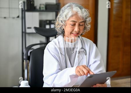 Felice e professionale medico asiatico anziano controllare il suo programma, rivedere la cartella clinica sul suo tablet digitale. Foto Stock
