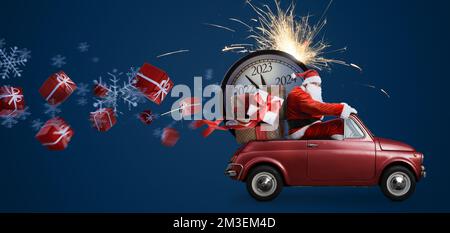 Natale sta arrivando. Babbo Natale su un'auto giocattolo che consegna regali per il nuovo anno 2023 e orologio per conto alla rovescia su sfondo blu con fuochi d'artificio Foto Stock