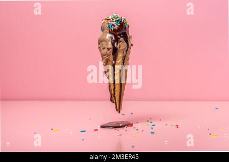 divertente concetto creativo di cono di wafer volante con gelato coperto, cosparso di spruzzi e versato con glassa di cioccolato, sfondo rosa Foto Stock