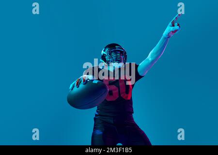 Sportivo, uomo forte, calciatore americano in uniforme sportiva e casco protettivo isolato su sfondo blu con luce al neon. Vista dal basso Foto Stock