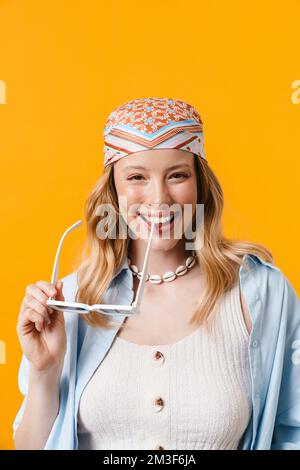 Giovane donna bionda sorridente alla macchina fotografica mentre si posa con occhiali da sole isolati su sfondo giallo Foto Stock