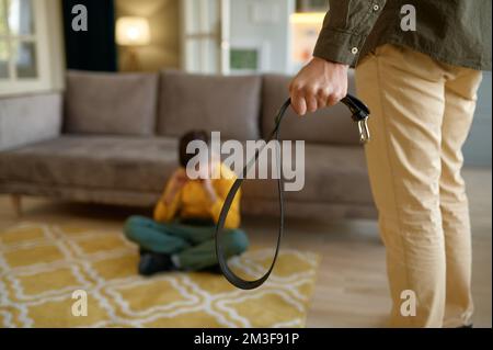 Primo piano del padre che tiene la cintura di pelle per punire il figlio piangente seduto sul pavimento Foto Stock