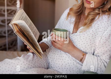 Primo piano donna incinta seduta a casa a letto, leggendo un libro e bevendo tè o acqua dalla tazza Foto Stock