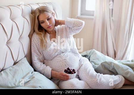 Donna incinta seduta sul letto e mangiare bacche di ciliegia. Modello biondo in pigiama sotto la coperta che tocca il collo Foto Stock