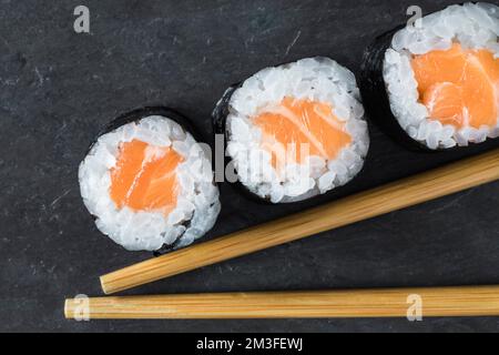 Vista dall'alto di 3 makis di salmone allineati su uno sfondo di pietra nera con bacchette. Orientale, cucina giapponese. Da asporto. Scatto macro. Foto Stock