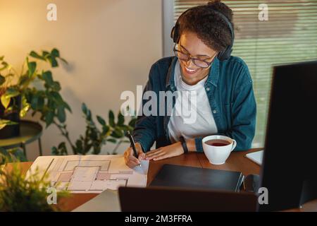 Sorridente Afro american donna interior designer in cuffie funziona con documenti in casa ufficio Foto Stock