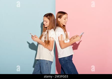 Chat in corso. Ragazze sorridenti, due sorelle amici in abiti casual utilizzando cellulari isolati su sfondo rosa-blu studio. Foto Stock