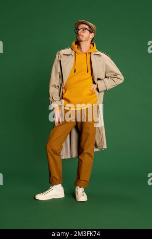 Ritratto di un giovane uomo alla moda in cappotto alla moda, berretto e felpa con cappuccio che posa isolato su sfondo verde. Stile autunnale Foto Stock