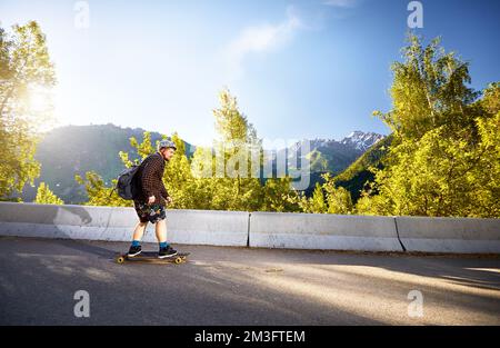 Uomo Skater nel casco cavalcare sulla strada di montagna al suo longboard vicino boschetto di legno di betulla e la cima della neve al Kazakhstan mattina d'estate Foto Stock