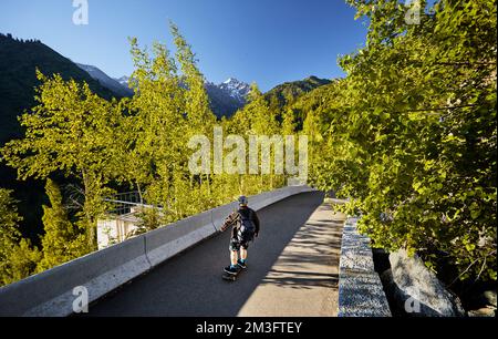 Uomo Skater nel casco cavalcare sulla strada di montagna al suo longboard vicino boschetto di legno di betulla e la cima della neve al Kazakhstan mattina d'estate Foto Stock