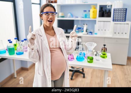 Giovane donna incinta che lavora al laboratorio scienziato stupito e sorpreso guardando in su e indicando con le dita e le braccia sollevate. Foto Stock