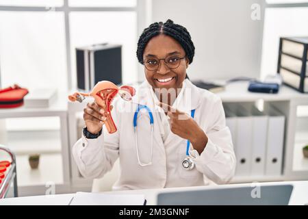 Donna afroamericana medico che tiene modello anatomico di organo genitale femminile sorridente felice puntamento con mano e dito Foto Stock