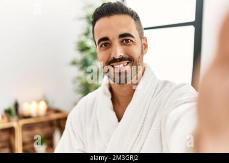 Giovane uomo ispanico che indossa un accappatoio facendo selfie con la macchina fotografica al centro di bellezza Foto Stock