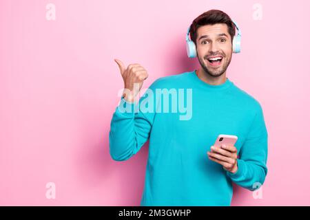 Foto di giovane ragazzo bearded indossare pullover blu ascoltare auricolari bluetooth dito punto mockup youtube musica app isolato su sfondo di colore rosa Foto Stock