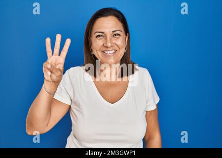 Donna matura ispanica in piedi su sfondo blu mostrando e indicando con le dita numero tre, mentre sorridendo fiducioso e felice. Foto Stock