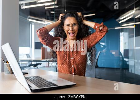 Donna d'affari arrabbiata urlando alla macchina fotografica, donna latino-americana che tiene la testa arrabbiata lavorando all'interno di un ufficio moderno utilizzando un computer portatile al lavoro. Foto Stock