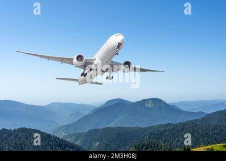 Un aereo di linea commerciale per passeggeri con carrello di atterraggio esteso sorvola il paesaggio collinare roccioso di montagna in una chiara giornata di sole Foto Stock