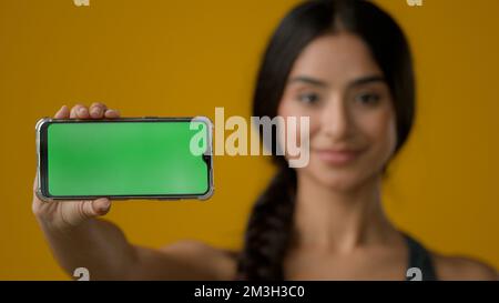 Primo piano etnico indiano donna sorridente ragazza in studio giallo tenendo telefono orizzontale mostrando verde vuoto schermo mobile di smartphone chroma key Foto Stock