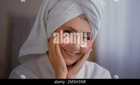 Ritratto positivo felice signora anziana con pelle antirughe hydrogel collagene patch sotto gli occhi sorridendo alla macchina fotografica vecchia 60s donna caucasica con asciugamano Foto Stock