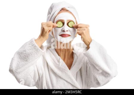 Donna in un accappatoio bianco con una maschera viso che mette su cetrioli sugli occhi isolati su sfondo bianco Foto Stock