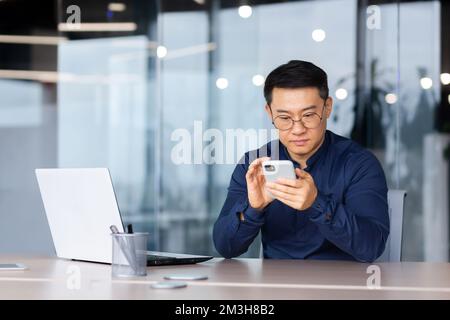 Primo piano di un serio e penoso uomo d'affari asiatico che utilizza il telefono, un uomo in ufficio a digitare un messaggio, navigare in Internet pagine e guardare video. Foto Stock