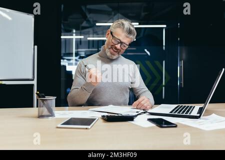 Uomo d'affari anziano felice seduto in ufficio al tavolo e la firma di un contratto di successo. fare un affare vantaggioso. Spettacoli felici con mano sì. Foto Stock