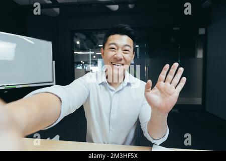 Ritratto di un giovane bell'uomo asiatico che prende un selfie, registra un video, parla durante una videochiamata, tiene in mano un telefono, agita e saluta la fotocamera, sorridendo. Foto Stock