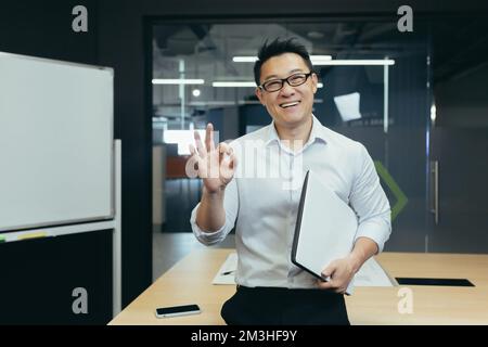 Ritratto di un giovane e bello studente asiatico, intern, assistente. Si alza con una cartella in ufficio, indossa occhiali, sorride, punta con le dita Foto Stock